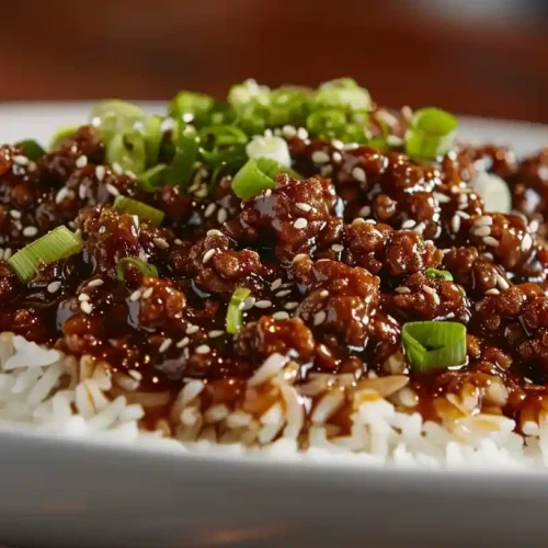 Mongolian ground beef served over steamed white rice, topped with green onions and sesame seeds, on a wooden table.