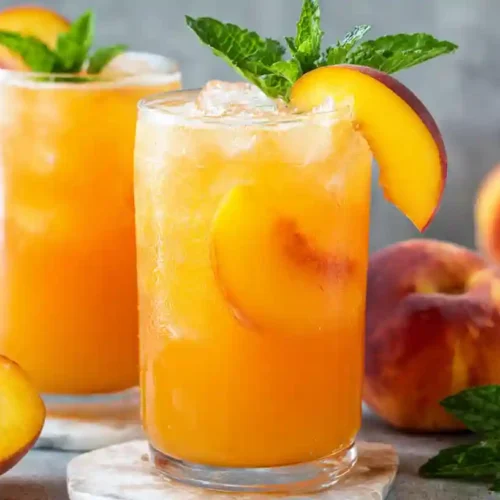 Glasses of easy peach lemonade with ice, peach slices, and mint sprigs, surrounded by whole and halved peaches on a gray background.