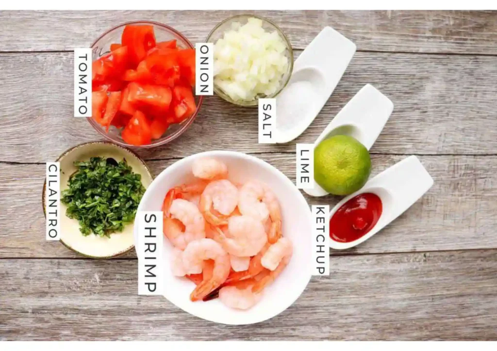 Classic ceviche ingredients laid out on a wooden surface: shrimp, diced tomato, onion, cilantro, lime, ketchup, and salt.